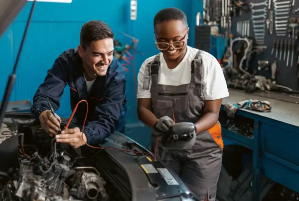 Two smiling auto technicians use a multimeter to test electrical components under the open hood of a car in a repair shop.