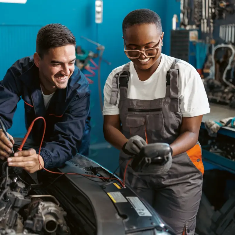 Two smiling auto technicians use a multimeter to test electrical components under the open hood of a car in a repair shop.