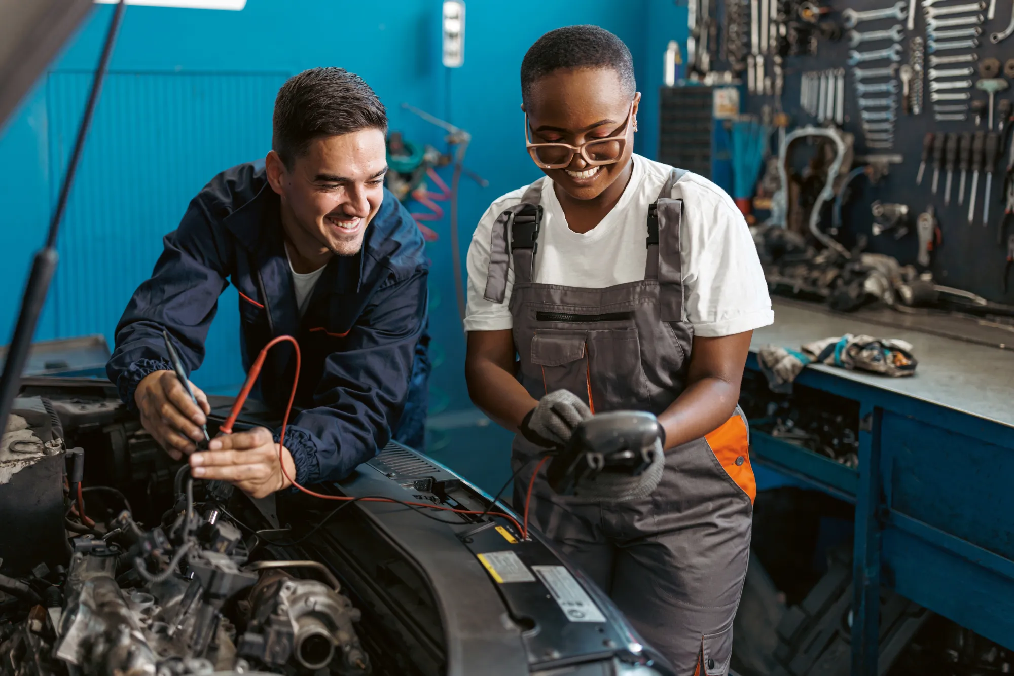 Two smiling auto technicians use a multimeter to test electrical components under the open hood of a car in a repair shop.