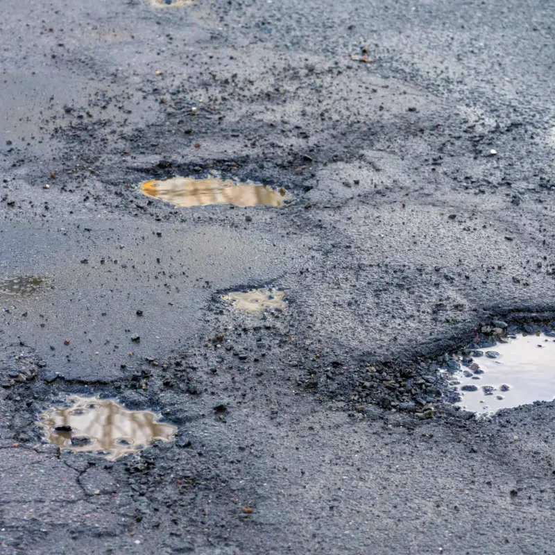 A cracked asphalt road surface with several potholes filled with muddy water and loose gravel around their edges.